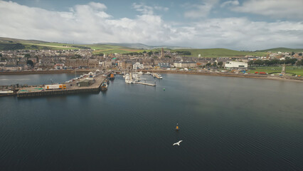Dock at pier town cityscape aerial. Seagull flight over ocean bay. Yachts, vessels at wharf. Old buildings architecture landmark at urban streets of Campbeltown city, Scotland. Cinematic seascape