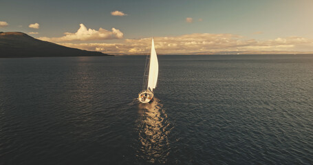 Aerial of yacht cruising in ocean bay waters at sun with clouds. White sail boat at open sea. Serene seascape with water transport. Summer vacation at sailboat. Cinematic relax and calm waterscape