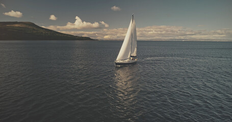 Sail boat at mountain ocean shore of Scotland Island of Arran aerial. Yacht sails with beautiful fluffy clouds sky at open sea. Sailboat cruise at summer sun shine day. Cinematic soft light drone shot