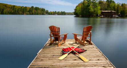 Two Adirondack chairs on a wooden dock face the serene blue waters of a Muskoka's lake. Canoe...