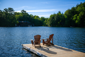 Soaking in the warmth of a sunny Muskoka morning, two Adirondack chairs rest peacefully on a wooden dock, overlooking the tranquil waters of the lake.