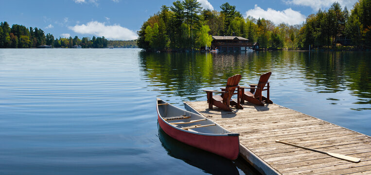 Two Adirondack chairs on a wooden dock overlook the tranquil blue waters of a Muskoka lake in Ontario, Canada. Nearby, a red canoe is tied to the pier, with life jackets and a paddle.