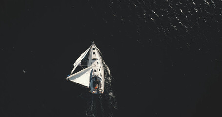 Top down of lone yacht sailing in open sea aerial. Black ocean and white sail boat at sun light reflection. Serene seascape with water transport at summer cruise scenery. Cinematic soft shot