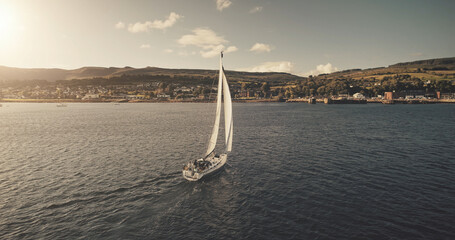 Aerial view of sun light on sail boat at ocean bay reflection. Highland sea coast with forest and mounts at bright sunlight. Nobody nature seascape at Brodick harbor with luxury yachts cinematic shot