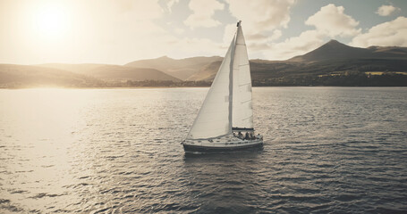 Fototapeta premium Sun seascape with white sailboat aerial. Mesmerezing landscape with sail boat at summer. Sunlight over sail boat at ocean bay. Sea shore of Arran Island with vessel. Serene water scenery drone shot