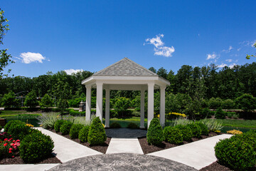 Fototapeta premium Gazebo in Lush Garden, Surrounded by Vibrant Flowers and Greenery Under a Clear Blue Sky, Walking Paths