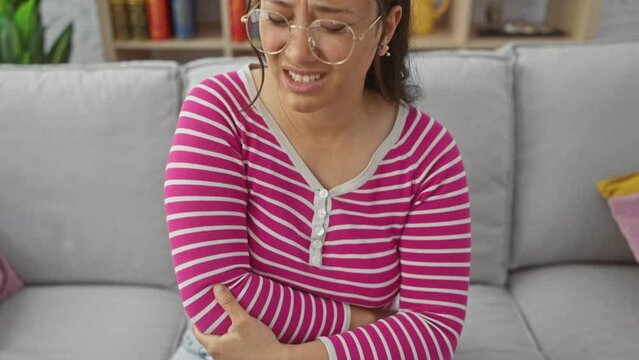 Pained woman with glasses sitting on a grey sofa indoors holding her elbow in discomfort.