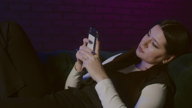 Bored adult young woman lying on divan at home and surfing news feed in smartphone. Young lady after job using phone on couch. Tired woman laying on divan and check her social medias