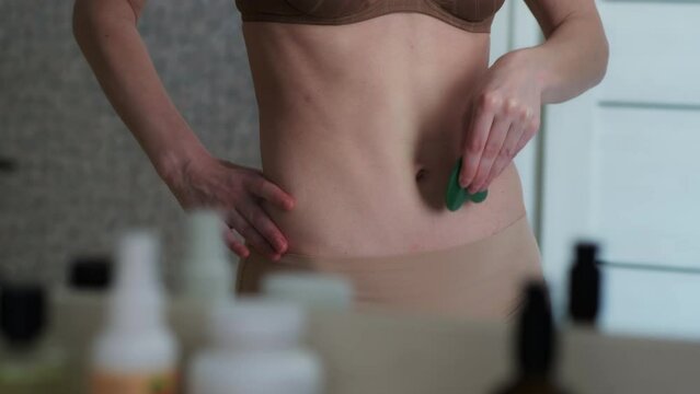 Adult woman in underwear making abdomen self-massage with stone scraper while standing in front of mirror. Using of lymphatic drainage massage techniques in home conditions.