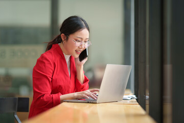 Asian businesswoman wearing glasses in a suit in the office is happy while using a smartphone to contact customers, online, video calls in business finance, marketing, taxes of the company.