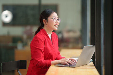 Confident Asian businesswoman in glasses sits on a chair in front of the counter, recording business financial goal plan online, analyzing, thinking, data entry on laptop of successful woman.