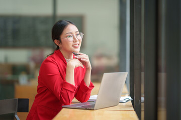 Confident Asian businesswoman in glasses sits on a chair in front of the counter, recording business financial goal plan online, analyzing, thinking, data entry on laptop of successful woman.