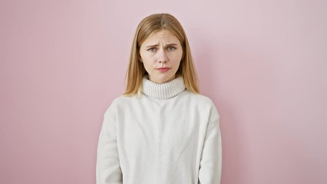 Disgruntled, beautiful blonde girl with a skeptic frown and nervous gaze stands isolated, grappling with an upset face over a problem. pink background augments the negative aura.