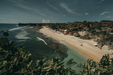 Amazing aerial Bali beach coast at ocean waves crashing to sand. Tropical paradise island of Indonesia with resting people and umbrellas on sandy coast. Cinematic exotic resort at shore of sea bay