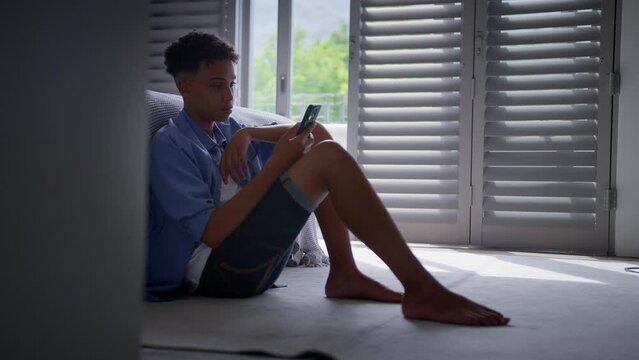 Anxious teenage boy sitting on bedroom floor at home with mobile phone concerned about bullying on social media - shot in slow motion