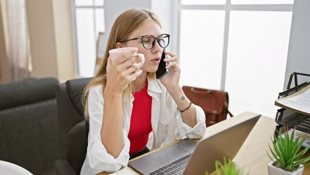 Blonde woman in glasses sipping coffee while talking on the phone at her office desk.