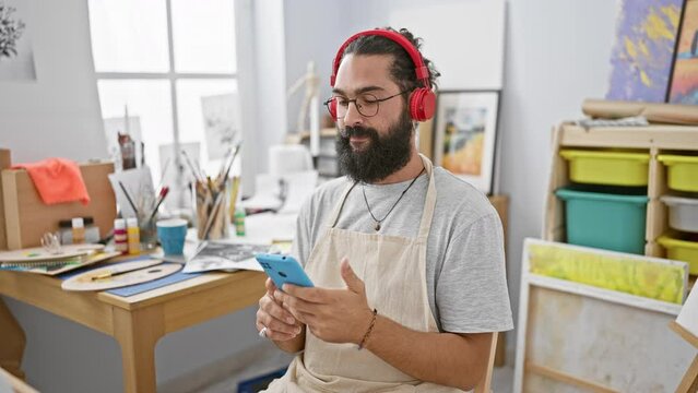 A bearded young man wearing headphones uses a smartphone in a creative art studio.