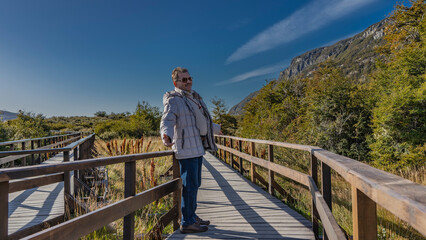 A man in a down jacket stands on a boardwalk by the railing. His arms are spread apart, smiling....