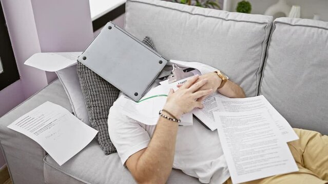Overwhelmed adult man surrounded by paperwork and laptop while sitting on a grey sofa indoors