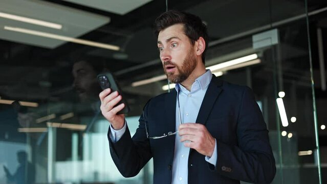 Side view of astonished caucasian male feeling shock while reading news on modern smartphone. Handsome businessman holding glasses in other hand and expressing emotions of surprise in modern office.