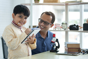Indian father teaches son to do exercises, boy wearing traditional clothes making note, Father's Day