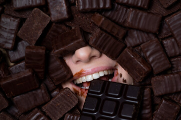 The face of a caucasian woman surrounded by chocolates. The girl eats a bar of chocolate.