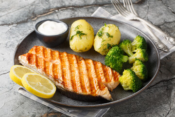 Dietary grilled salmon with boiled potatoes, broccoli, lemon, herbs and cream sauce close-up in a plate on the table. Horizontal