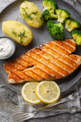 Portion of grilled salmon with boiled potatoes, broccoli, lemon, herbs and cream sauce close-up in a plate on the table. Vertical top view from above