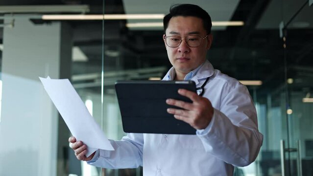 Male therapist holding documents and tablet while comparing information on paper and online. Man in uniform with stethoscope using digital gadget for efficient work with client database in clinic.