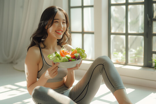 Beautiful fit woman eating healthy salad after fitness workout, Young Asian woman enjoys a fresh salad, embodying a picture of health and happiness in her bright home.