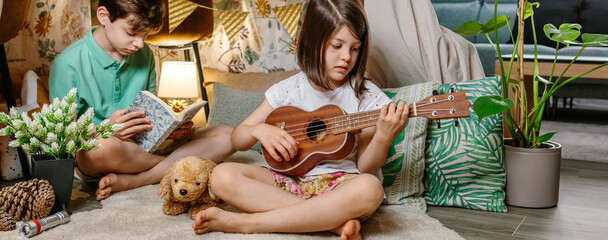 Banner of little girl playing ukulele while boy reading book on handmade teepee in living room. Children having fun in diy shelter tent in their house. Vacation camping at home or staycation concept.