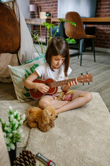 Little girl playing ukulele sitting on carpet next to handmade shelter tent in living room. Concentrated child practicing music next to diy teepee. Vacation camping at home or staycation concept.
