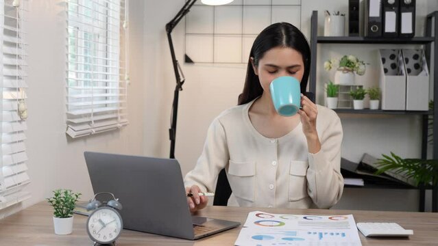 Confident Asian businesswoman working on real estate project, finance, accounting and drinking coffee while working, relaxing, taking breaks, using phone, laptop in home office.