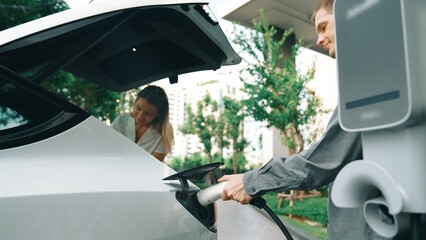 Young couple travel with EV electric car charging at green sustainable home in summer shows urban...