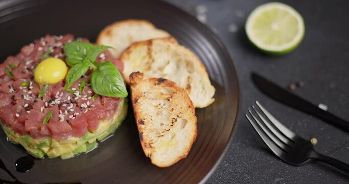 Tuna and avocado tartare with sesame seeds, capers and egg yolk on a dark ceramic plate with baguette bread crouton chips