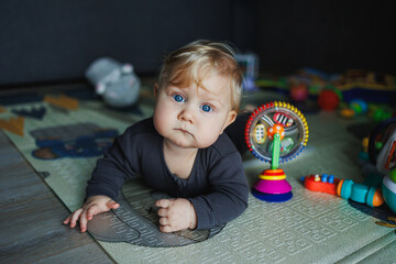 A 7-month-old baby is crawling on a rug. The child holds his head. A baby learns to crawl on the floor. A child in a gray romper.