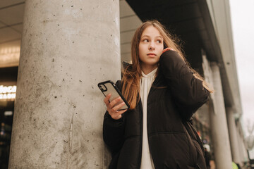 Half length portrait of girl in casual wear holding smartphone for blogging in the city in the autumn-winter season. Teen surfing the internet outdoors - 782856554