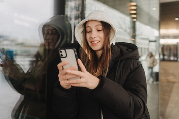 Half length portrait of girl in casual wear holding smartphone for blogging in the city in the autumn-winter season. Teen surfing the internet outdoors - 782857320