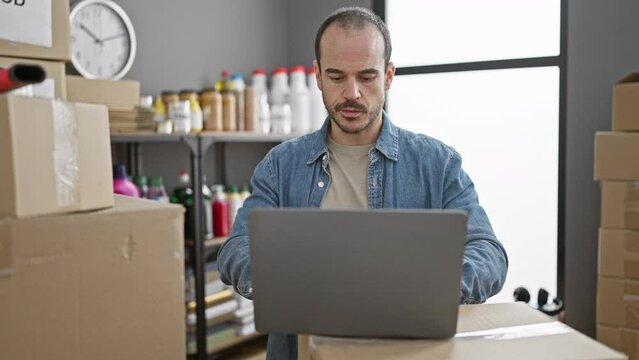 Bald man with beard in warehouse using laptop surrounded by cardboard boxes and donations