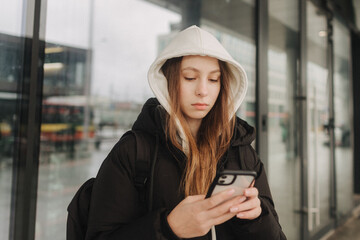 Half length portrait of girl in casual wear holding smartphone for blogging in the city in the autumn-winter season. Teen surfing the internet outdoors - 782857522