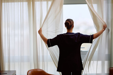 Rear view of chambermaid opening curtains in hotel room.