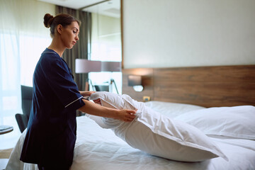 Young female housekeeper making bed in hotel room.