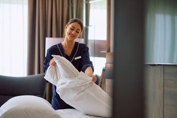 Smiling maid changing bedding in hotel room. - obrazy, fototapety, plakaty