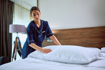 Young chambermaid making bed while working in  hotel. - obrazy, fototapety, plakaty
