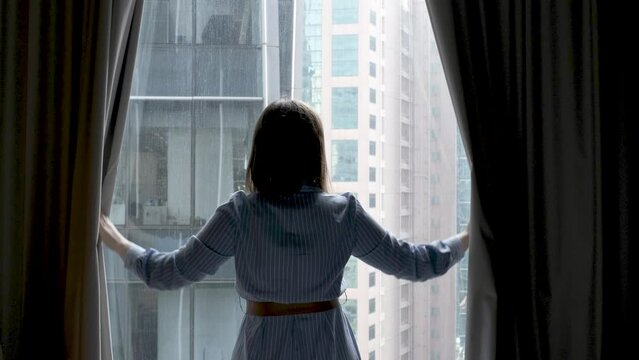 A woman in blue dress opens curtains and looks out the window.