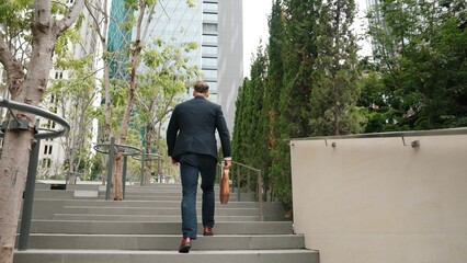 Back view of caucasian manager with formal suit walking up stairs while holding his suitcase in green city. Professional business man going to workiplace while climb stair in eco urban city. Urbane..