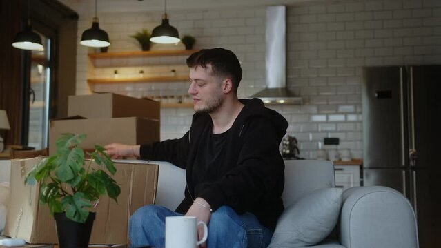 Unhappy young man feeling stressed moving into new apartment or moving out of house, sitting on sofa with carton boxes, tired of long renovation relocation processes