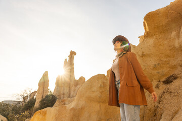 A woman wearing a brown jacket and hat stands on a rocky hillside. The sun is shining brightly, casting a warm glow on the scene. The woman is enjoying the beautiful weather and the stunning landscape - Powered by Adobe