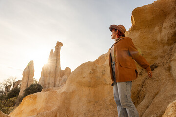 A woman wearing a brown jacket and jeans stands on a rocky hillside. The sun is shining brightly,...