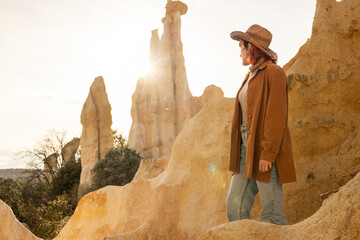 A woman wearing a brown jacket and a straw hat stands on a rocky hillside. The sun is shining...